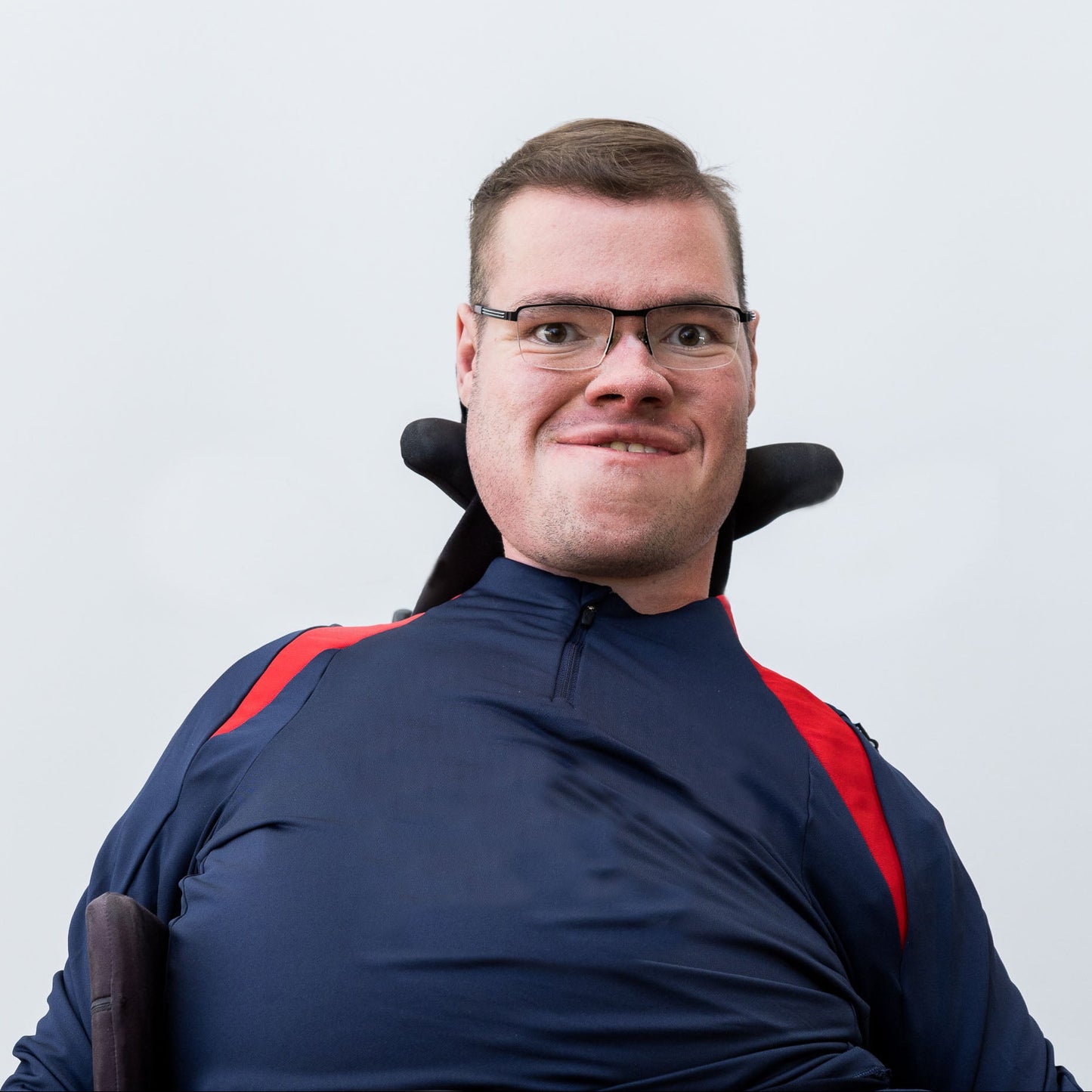 Man in a wheelchair wearing glasses and a dark blue jacket with red accents against a light gray background