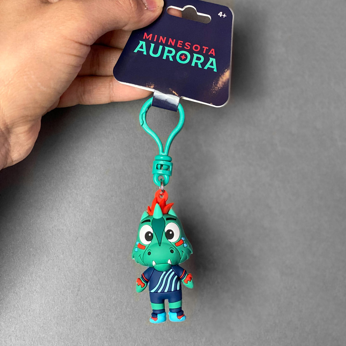 Plastic keychain of the Aurora mascot, Rory the Aurorasaurus, in a navy blue soccer jersey