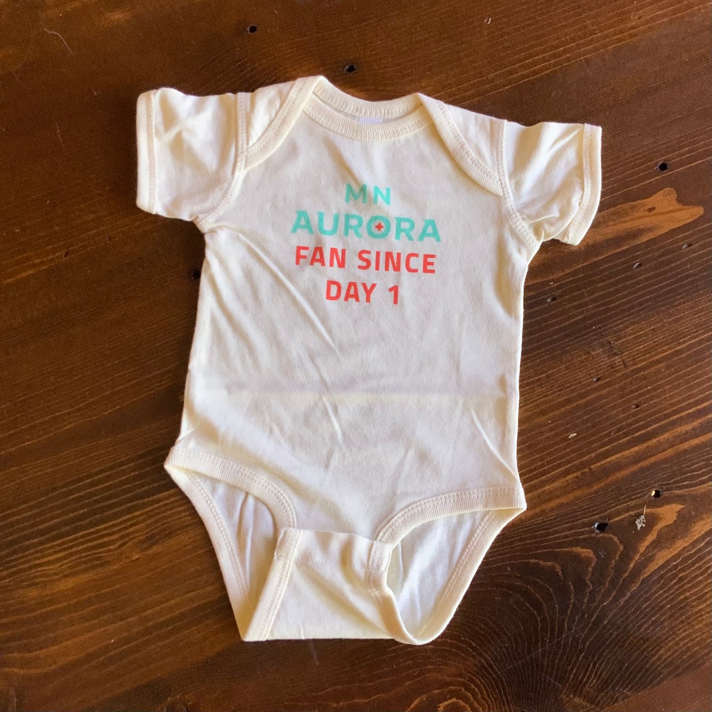 White onesie with the words "MN Aurora Fan Since Day 1" in teal and orange