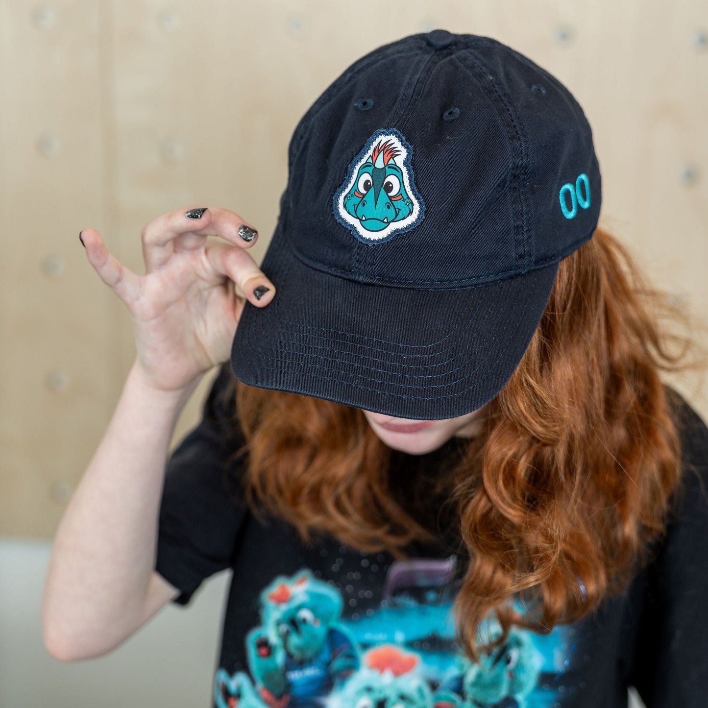 Navy blue hat with a patch of the Aurora mascot, Rory the Aurorasaurus, featuring her name and number "00" on the sides embroidered in teal