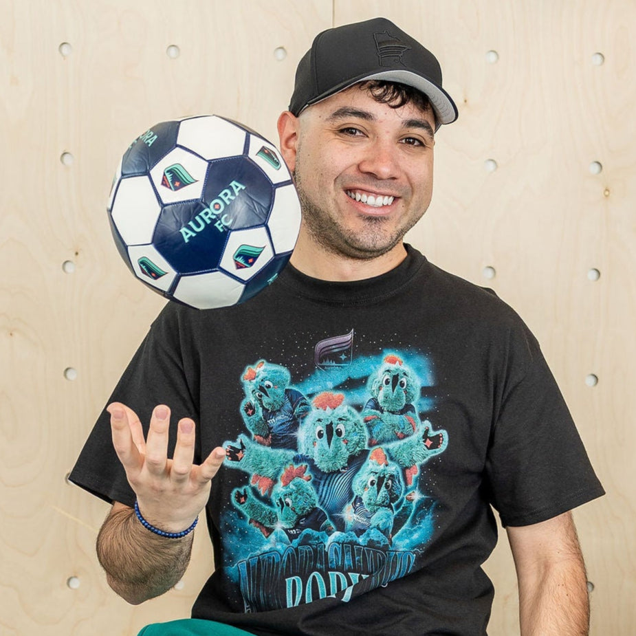 Black short-sleeved shirt featuring a collage of the Aurora mascot, Rory the Aurorasaurus, with the aurora borealis in the background