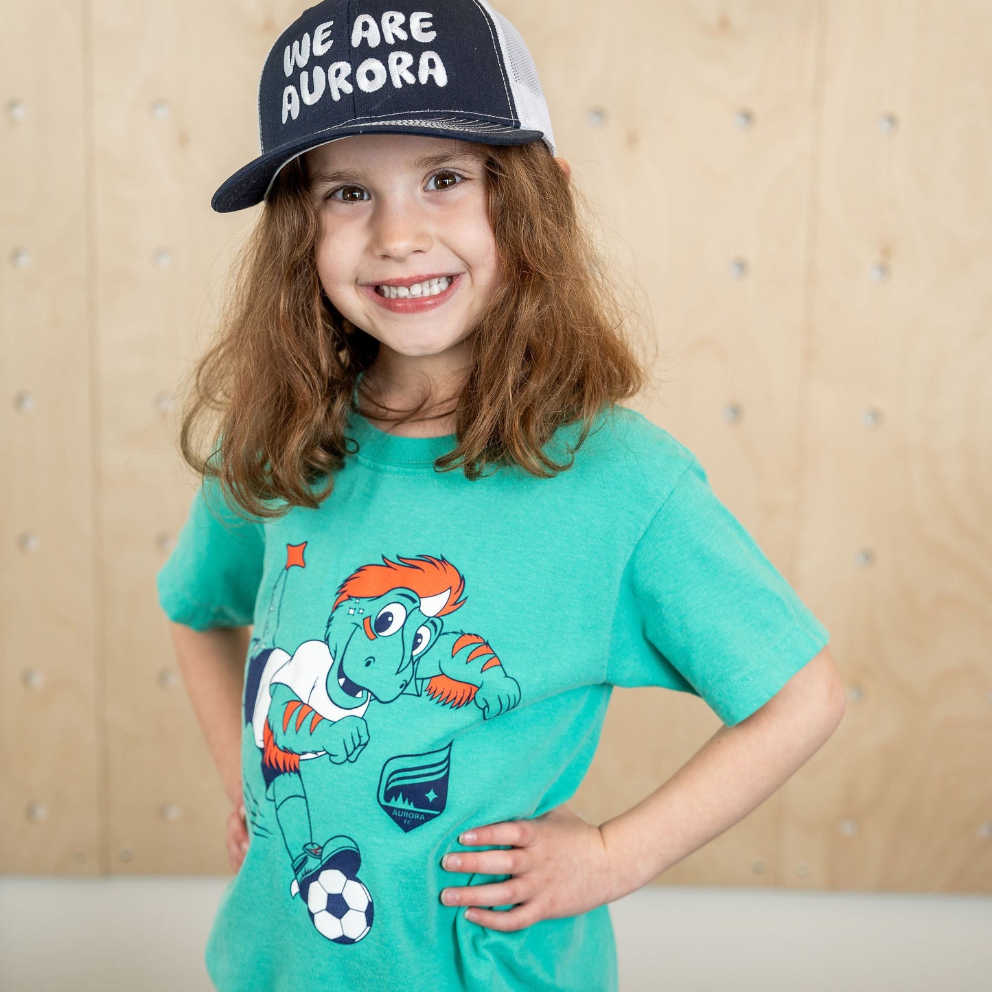 Teal short-sleeved shirt featuring a picture of the Aurora mascot, Rory the Aurorasaurus, running while dribbling a soccer ball