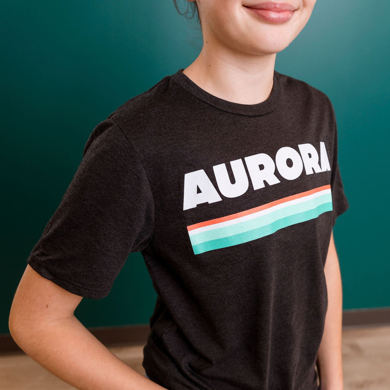 Person wearing a black t-shirt with 'AURORA' printed on it against a teal wall.