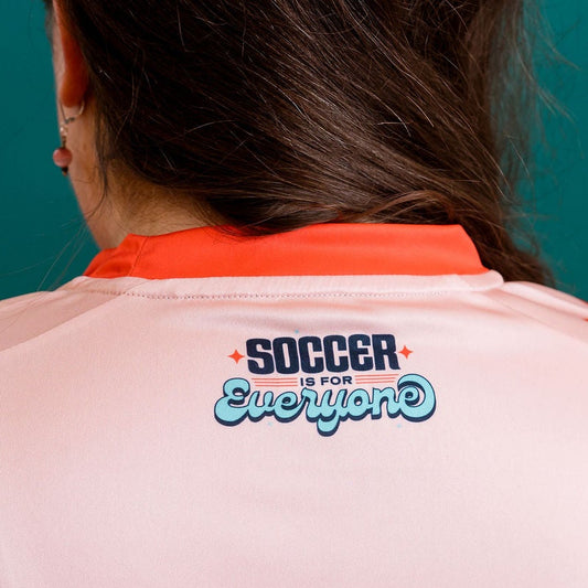 Person wearing a salmon colored soccer jersey with red accents against a teal wall.