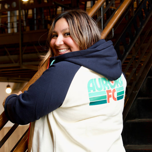 A woman smiling and looking back over her shoulder while wearing a cream and navy raglan hoodie with a green and orange 'AURORA FC' graphic on the back, standing in an indoor setting with a staircase.