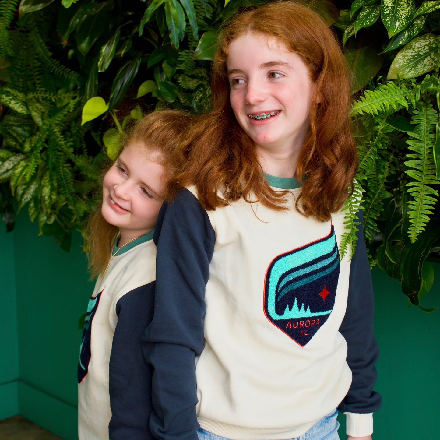 Two young girls with red hair wearing matching cream and navy blue sweatshirts with a colorful shield graphic on the front, standing in front of a green leaf wall.