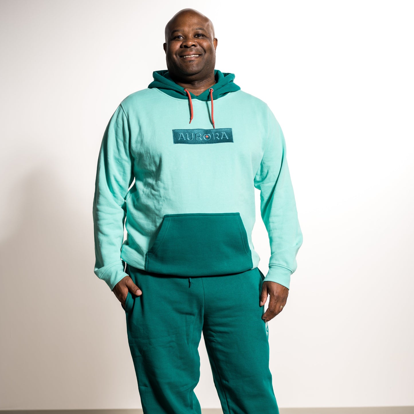 Teal hoodie with the front pocket and hood in dark teal and the rest in light teal with the word "Aurora" in a dark teal box and orange drawstrings