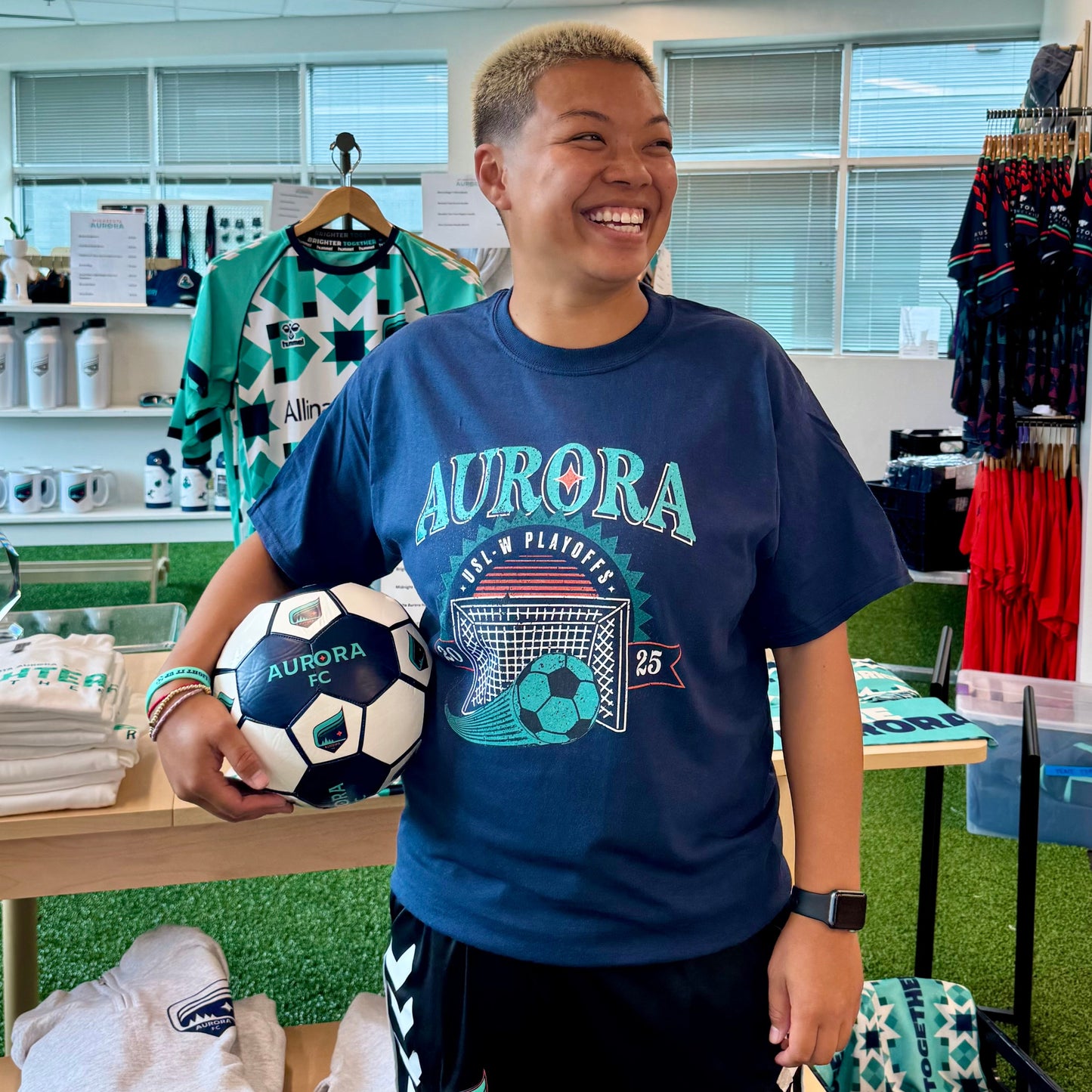 Person wearing a blue 'Aurora' t-shirt holding a soccer ball in a store setting