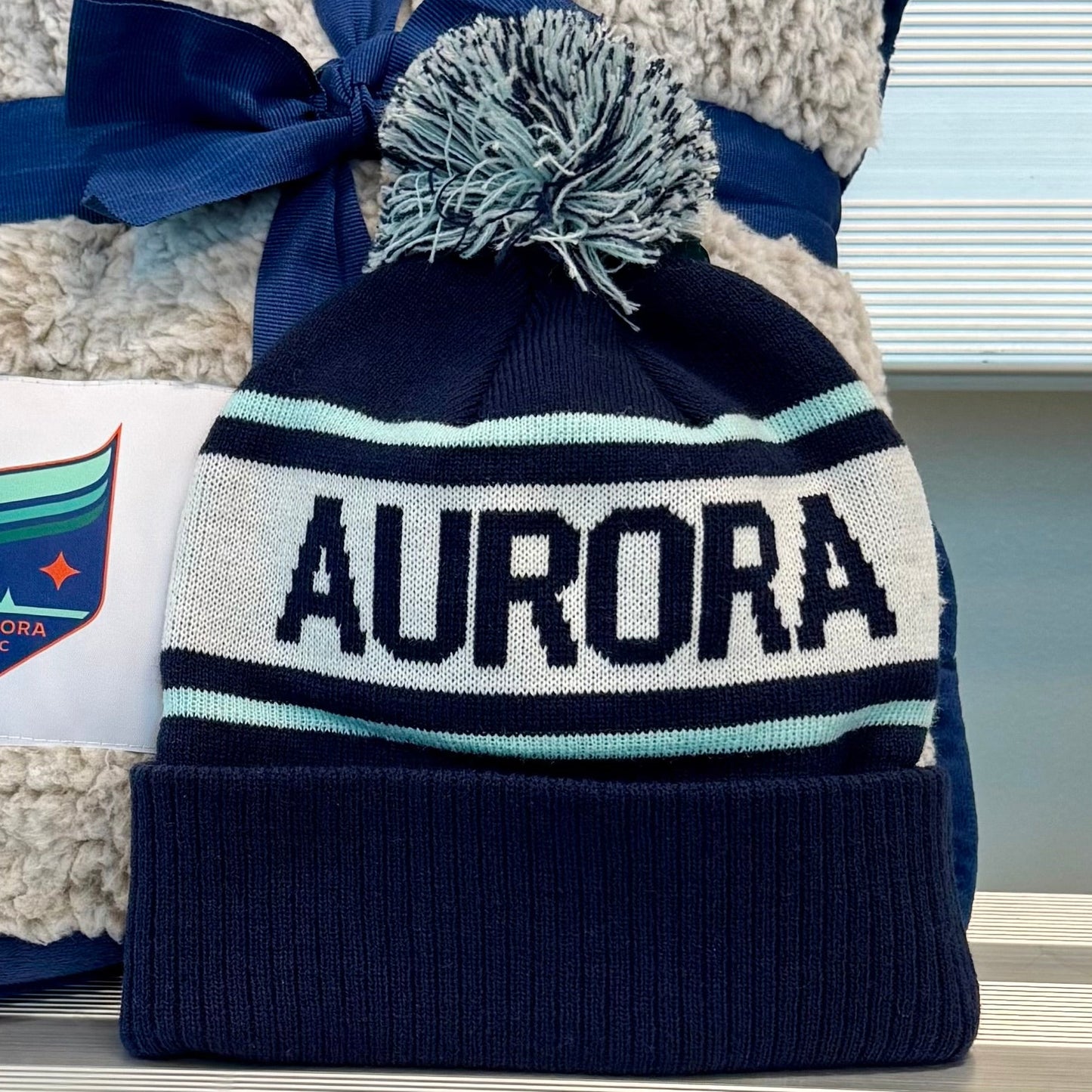 Navy and white beanie with pom-pom and 'Aurora' text, paired with a matching blanket on a metallic surface.