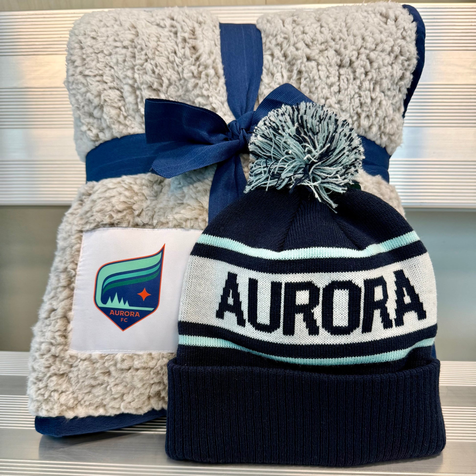 Navy beanie with 'Aurora' text and pom-pom, tied to a plush gray blanket with a visible brand logo.