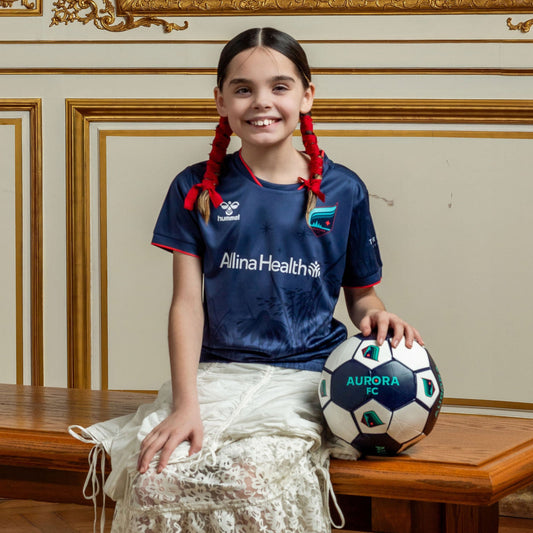 Young girl with braided hair sitting on a wooden bench holding a soccer ball in an ornate room.
