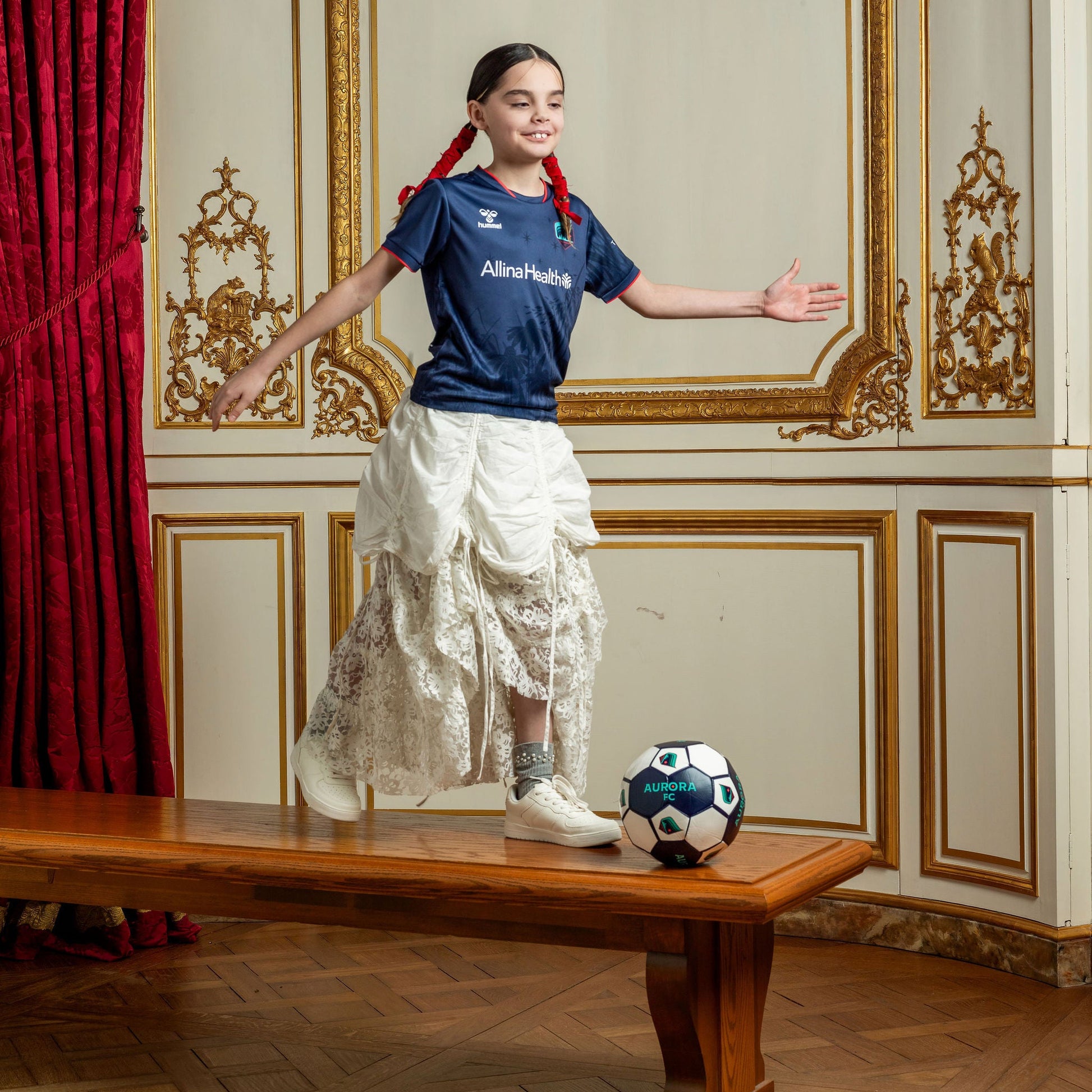Young girl in navy soccer jersey and a formal skirt with a soccer ball in an ornate room