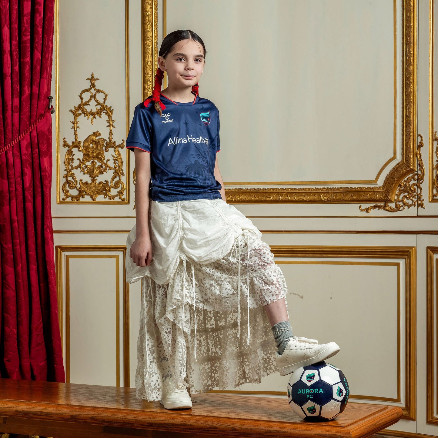 Young girl in a formal outfit with a soccer ball on a wooden table in an ornate room.