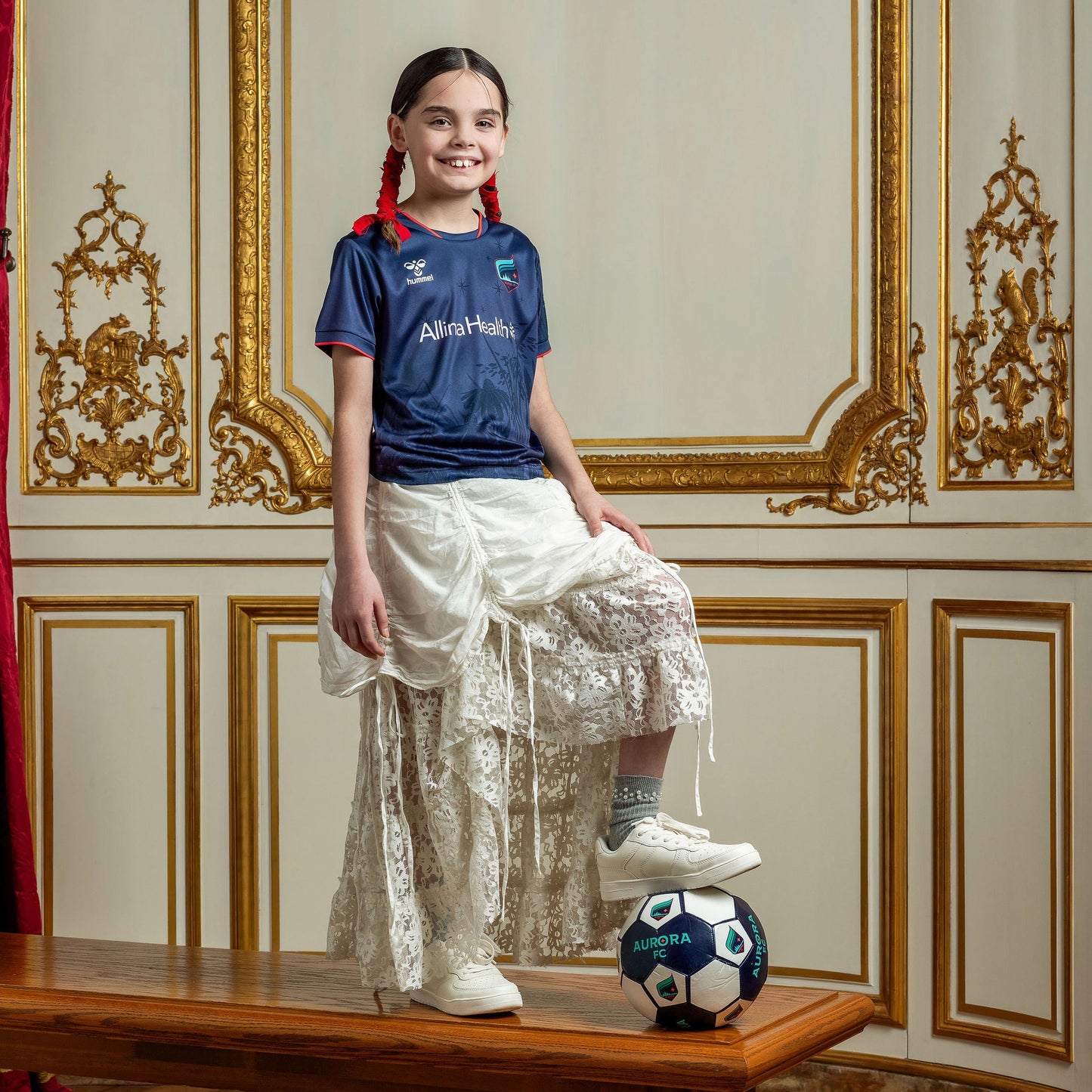 Young girl in a blue shirt and white skirt standing on a wooden bench with a soccer ball in an ornate room.