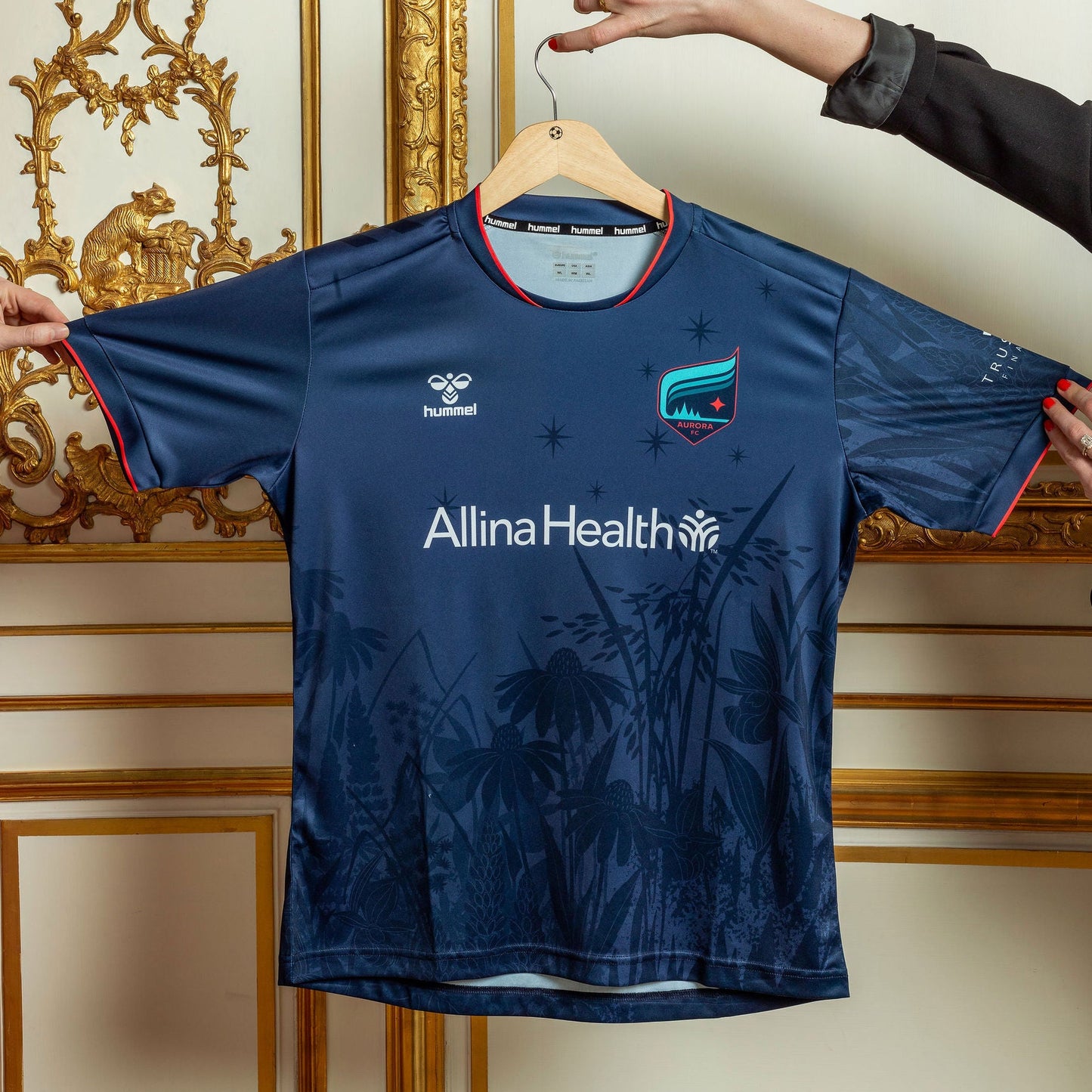 Navy blue sports jersey with 'Allina Health' logo on a hanger against a decorative wall.