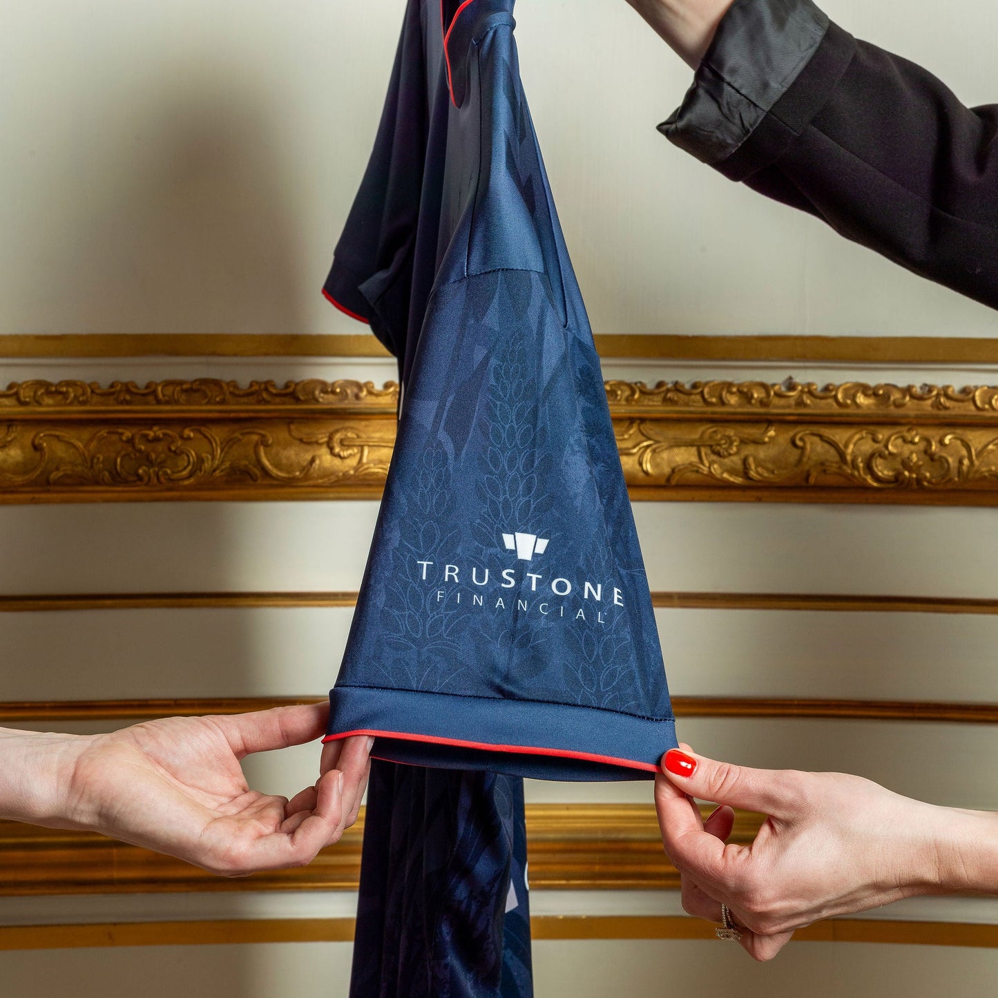 Navy blue jersey with TruStone Financial logo held by two hands against a decorative wall.