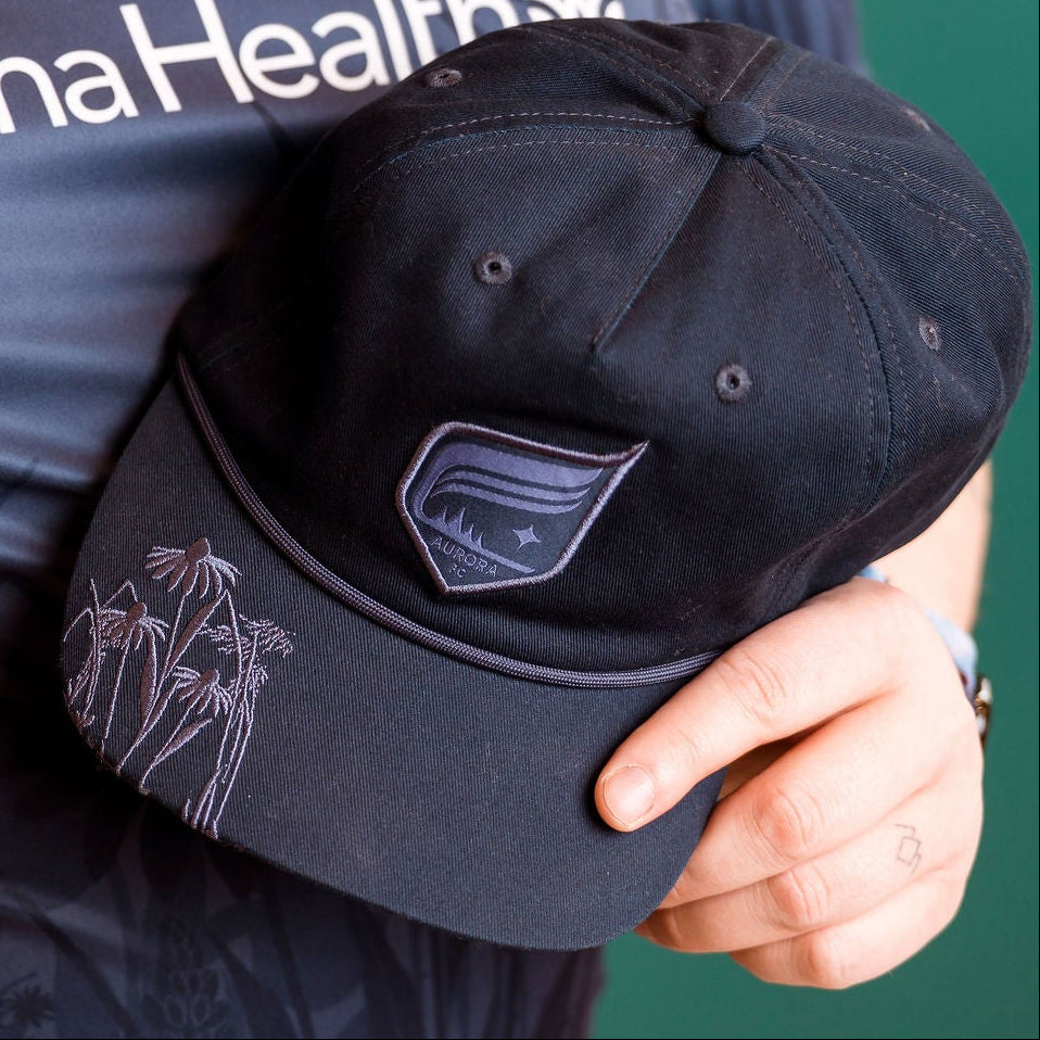 Person wearing a navy cap with a logo, holding it in front of an Allina Health shirt.