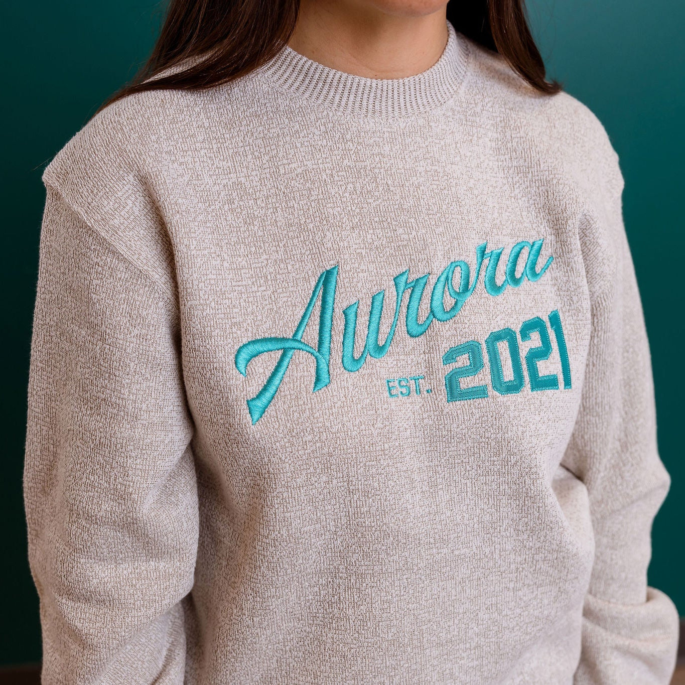 Person wearing a beige sweater with 'Aurora est. 2021' text in teal on a dark background