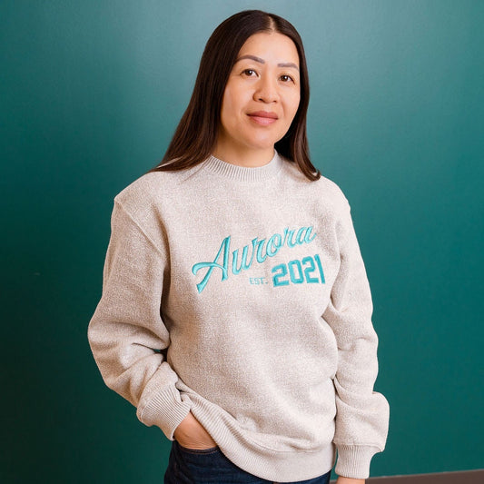 Woman wearing a light tan sweatshirt with text standing against a teal wall.