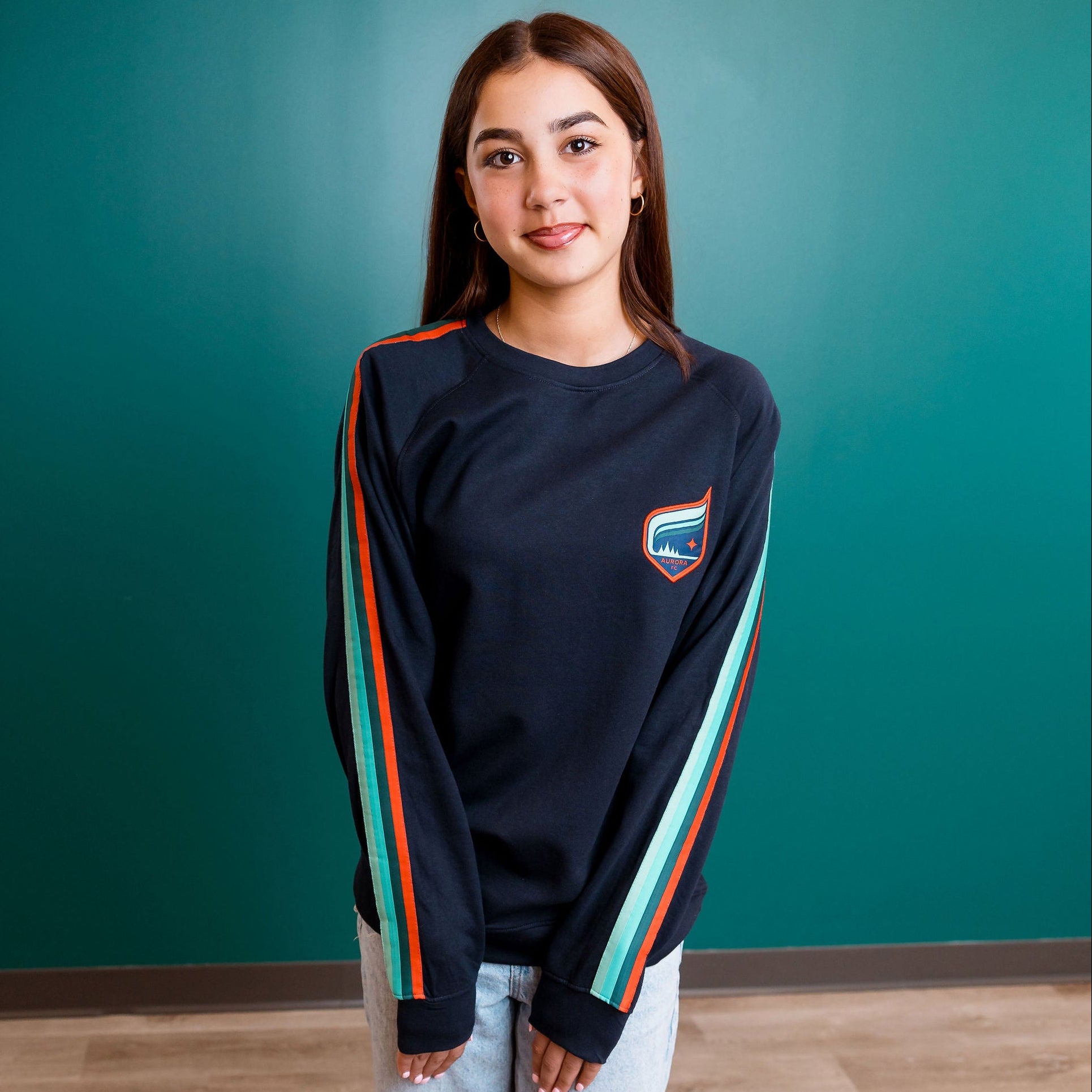 Person wearing a dark blue sweatshirt with colorful stripes and light blue jeans against a teal wall.