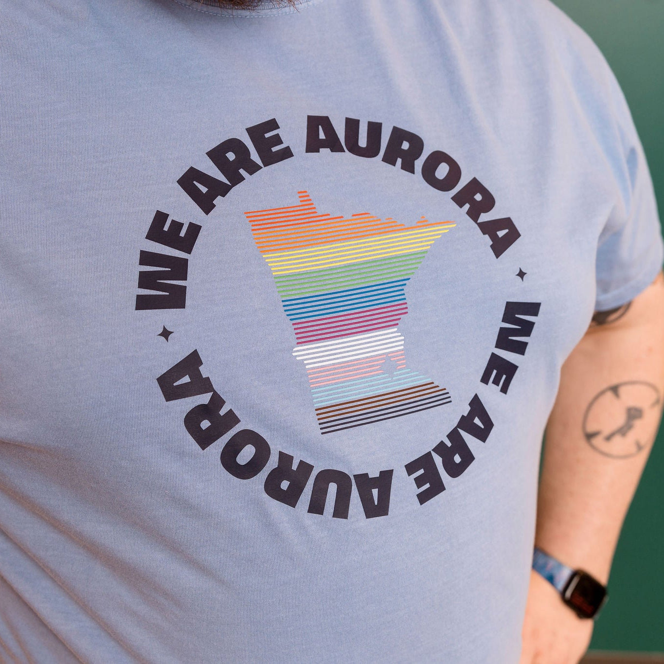 Person wearing a light blue t-shirt with 'We Are Aurora' design featuring a rainbow flag.