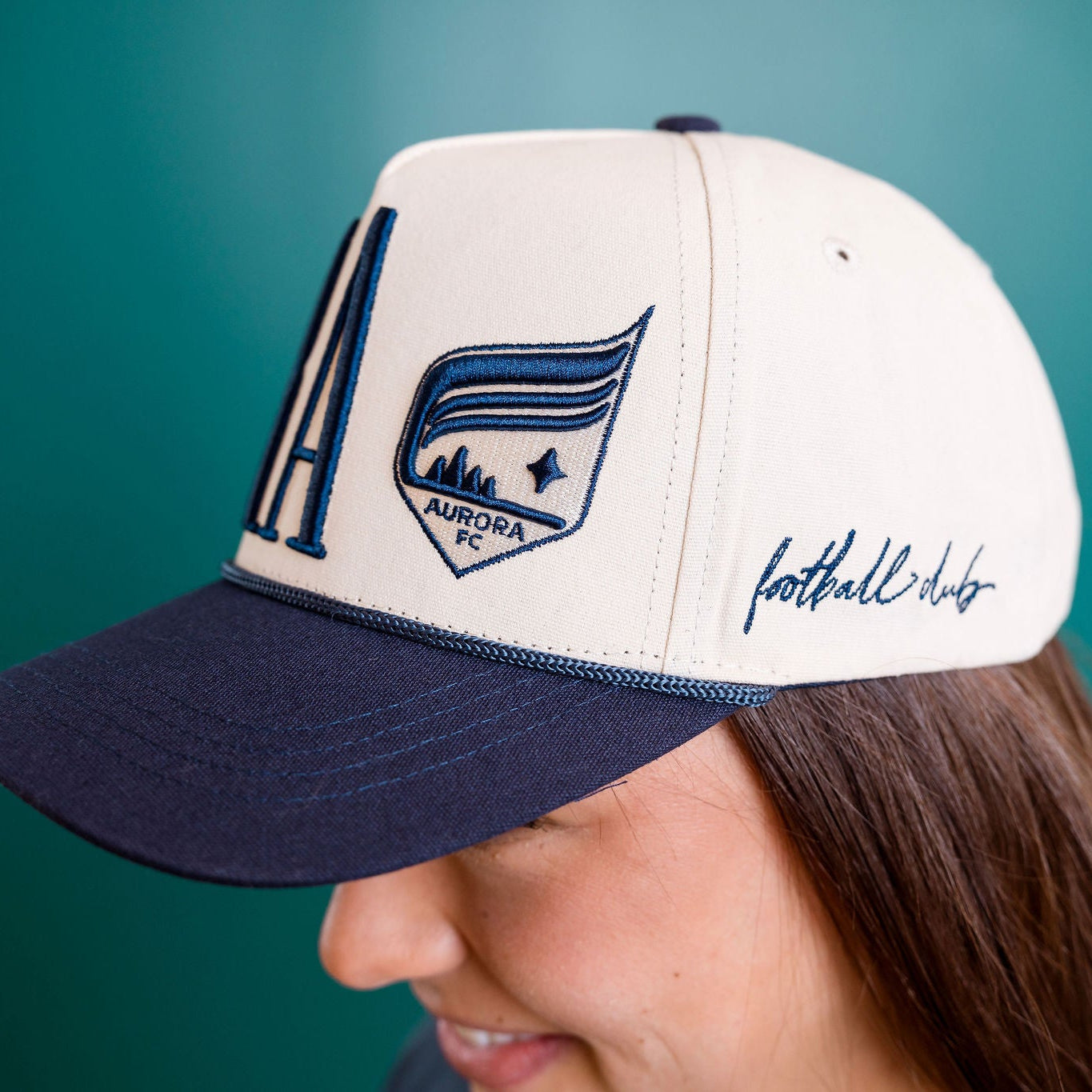 Person wearing a white and navy cap with 'Aurora Football Club' logo on a teal background