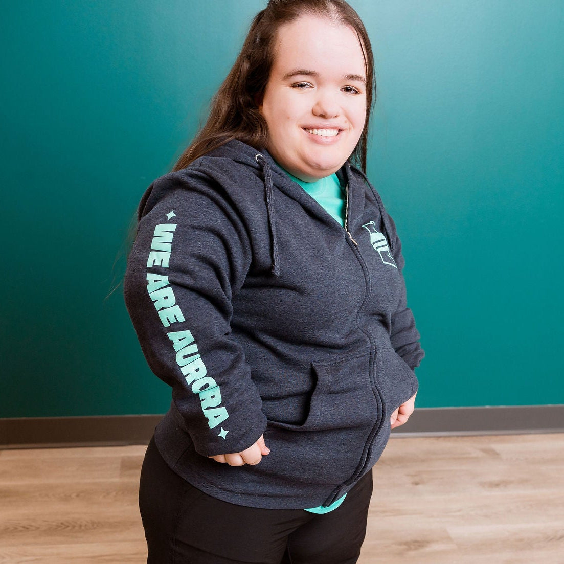 Person wearing a dark hoodie with text, standing indoors against a teal wall.