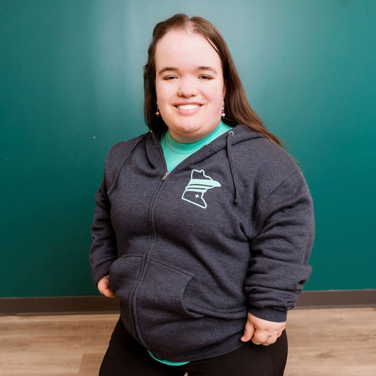 Person wearing a dark gray hoodie with a logo in front of a teal wall.