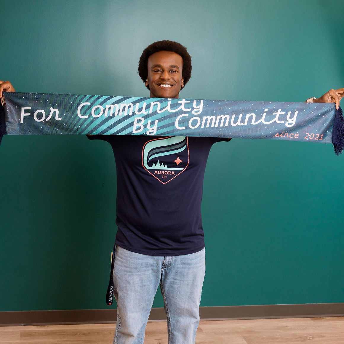 Person holding a scarf with "for community by community" text and a logo in an indoor setting