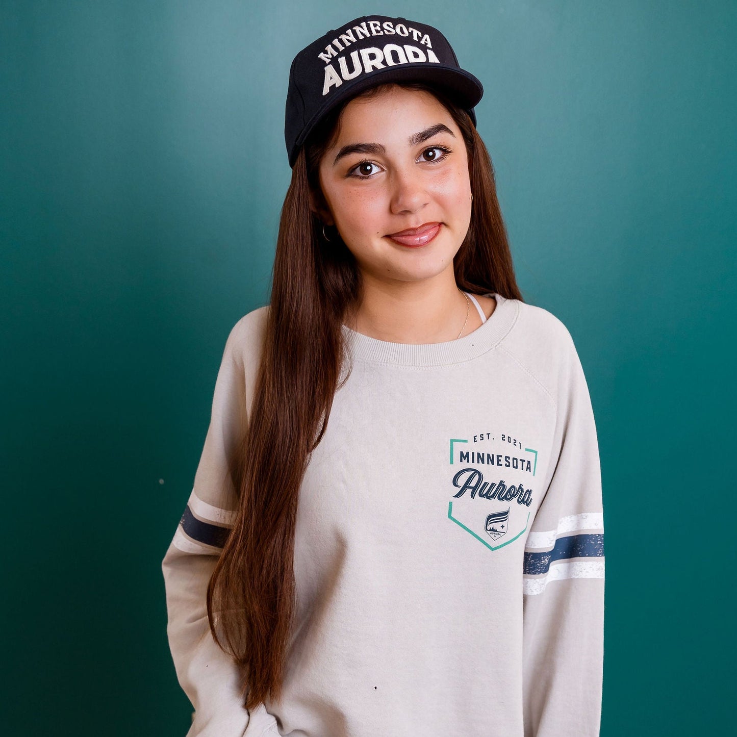 Person wearing a beige sweatshirt and cap against a teal wall