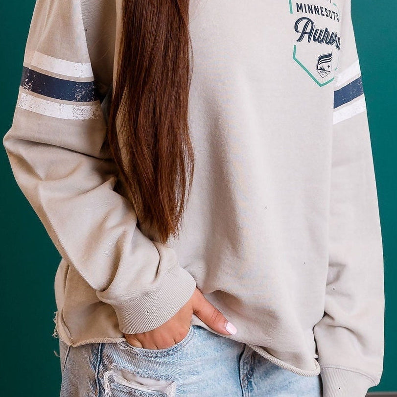 Person wearing a beige cap and light-colored sweatshirt with ripped jeans against a teal wall.