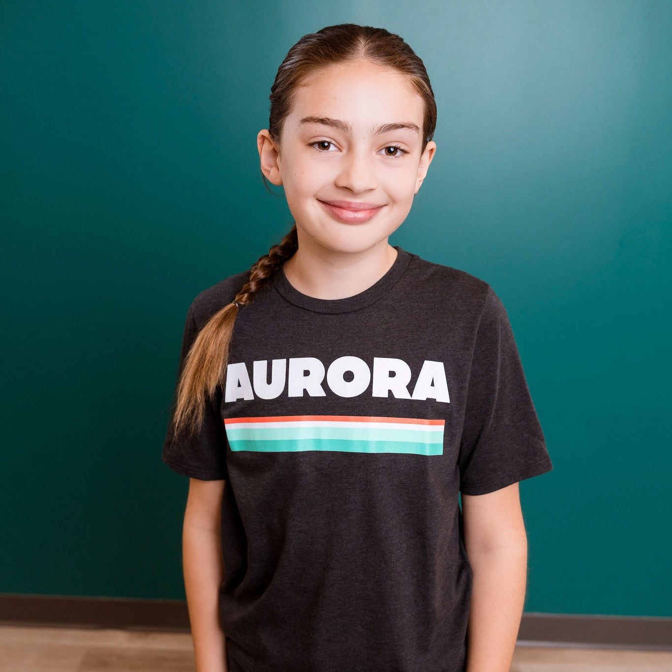 Young girl wearing a black t-shirt with 'AURORA' printed on it against a teal wall.