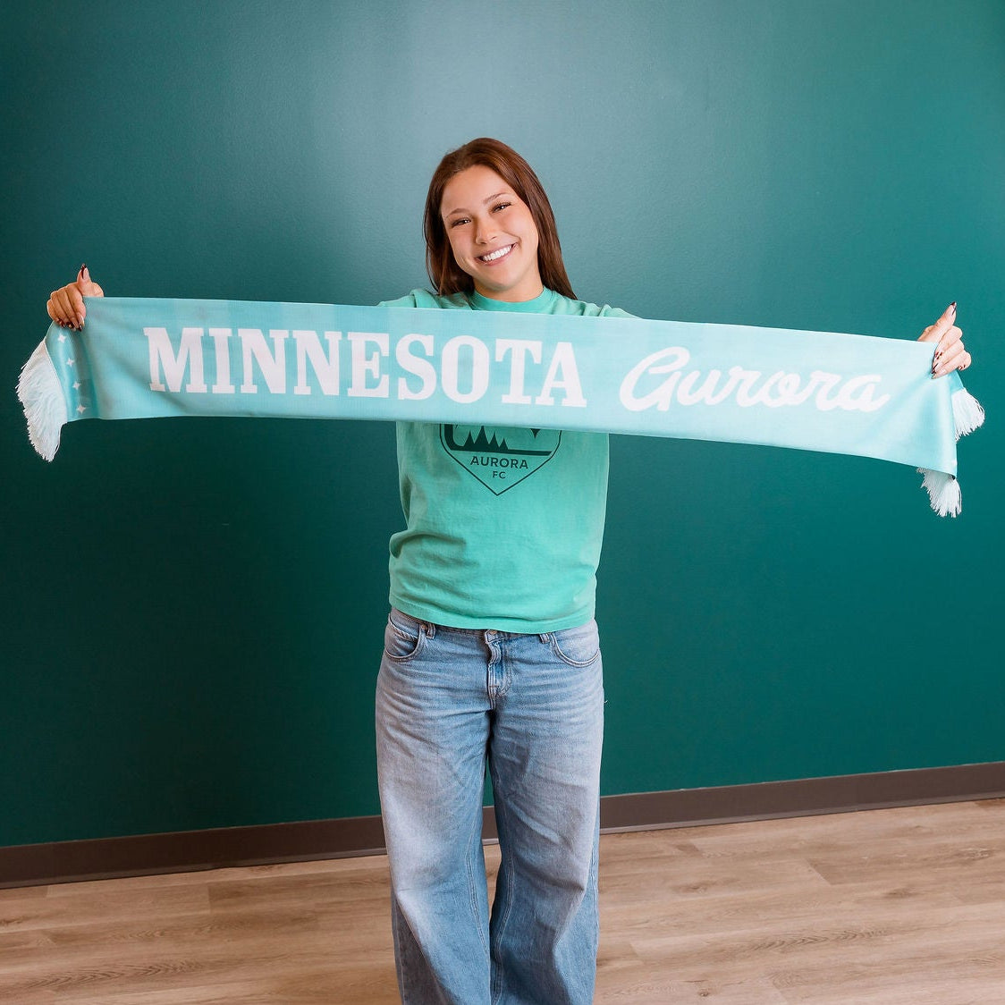 Person holding a 'Minnesota' scarf in front of a teal wall