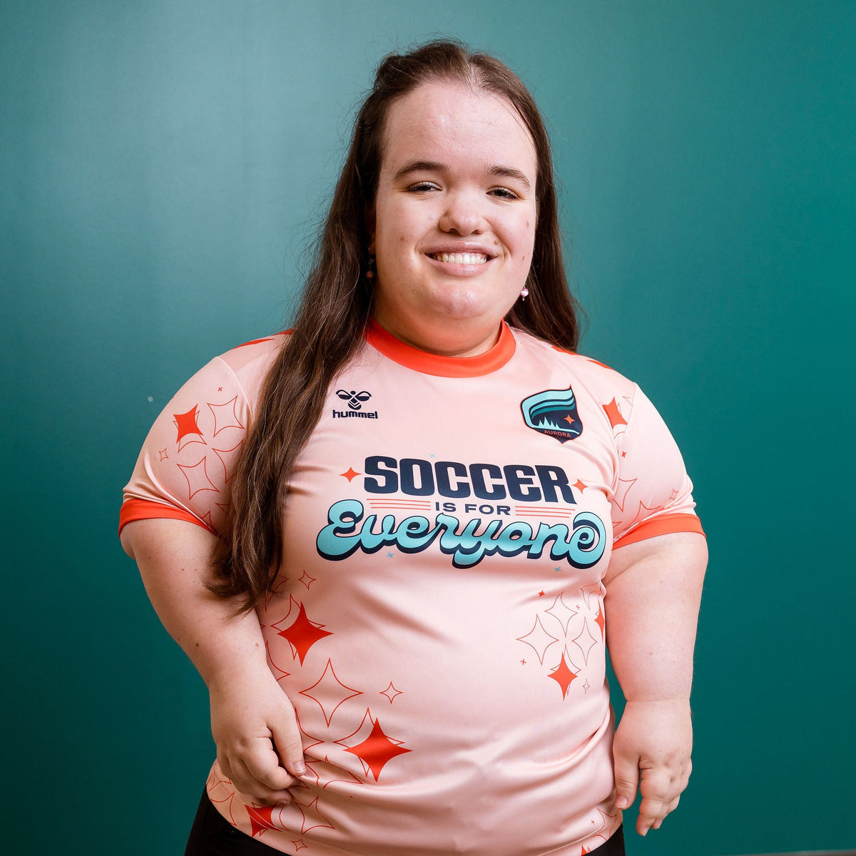 Person wearing a pink 'Soccer is for Everyone' jersey against a teal wall.