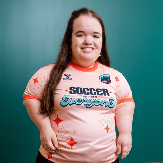 Person wearing a pink 'Soccer is for Everyone' jersey against a teal wall.