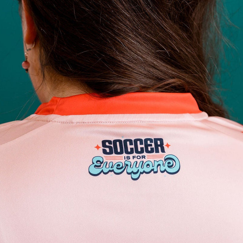 Person wearing a salmon colored soccer jersey with red accents against a teal wall.