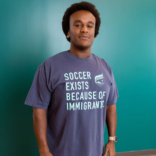 Person wearing a t-shirt with a message about soccer and immigrants against a teal wall.