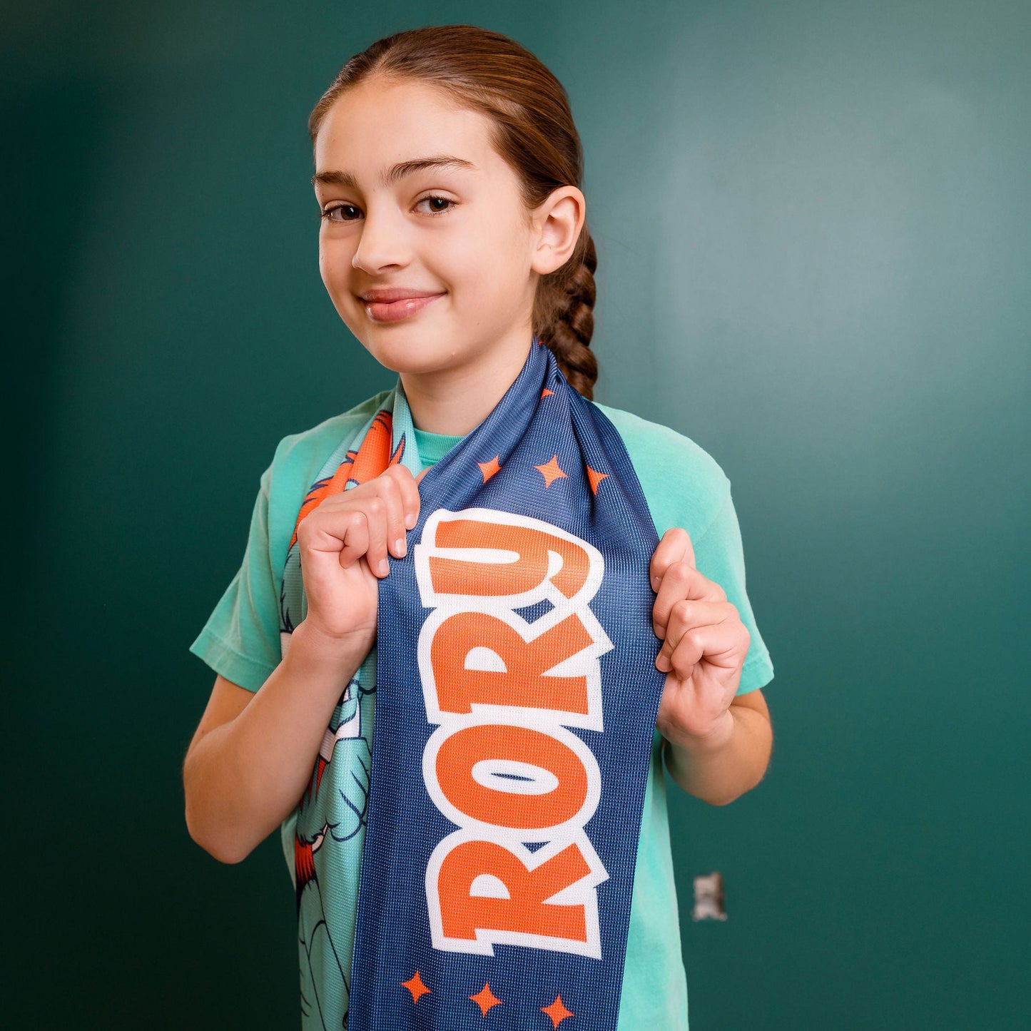 Young girl holding a blue scarf with 'RORY' printed on it against a green wall.