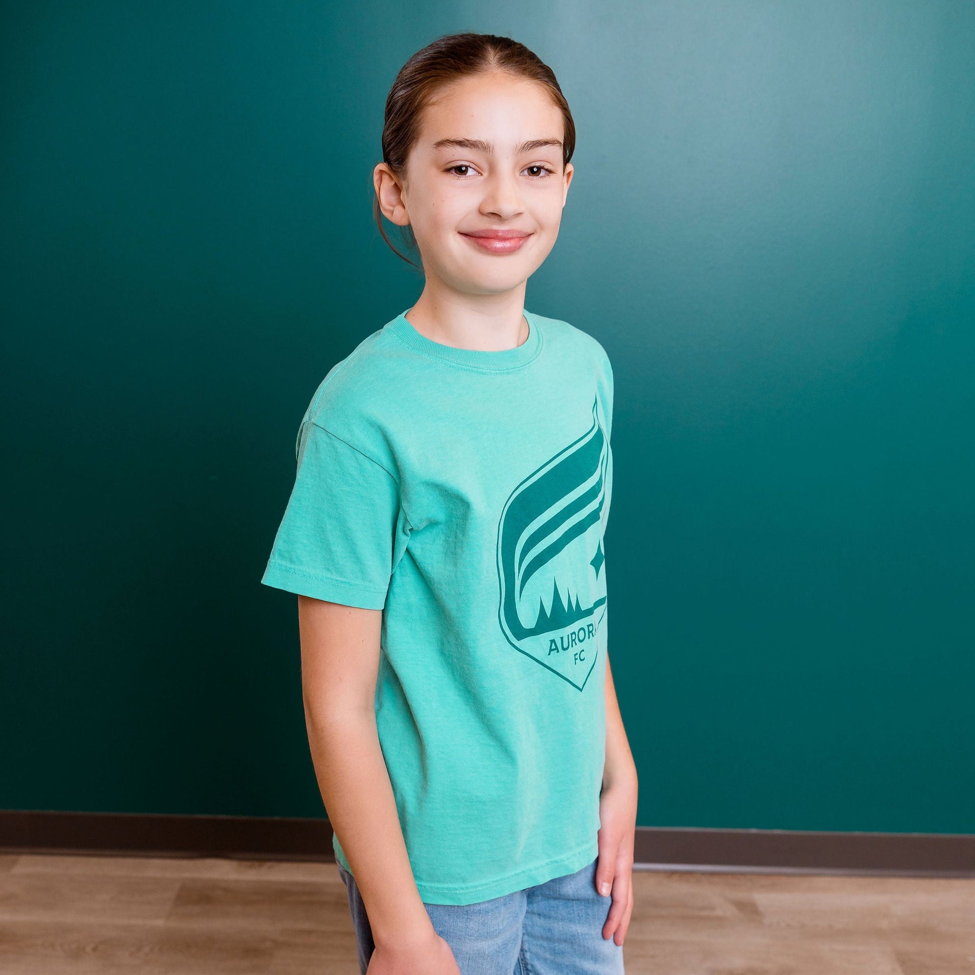 Person wearing a teal t-shirt with a logo against a teal wall.