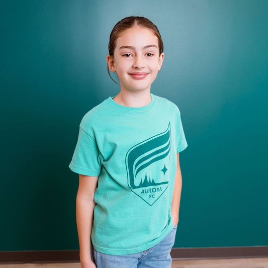 Young girl wearing a teal t-shirt with a logo against a teal wall.