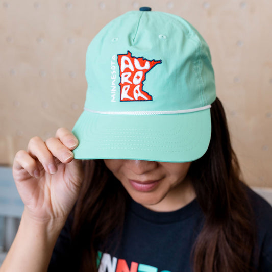 Person wearing a light teal cap with a logo, covering their face, against a neutral background.