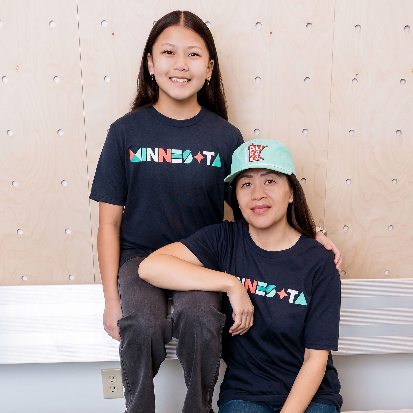 Two people sitting on a bench wearing 'MINNESOTA' t-shirts against a textured wall.