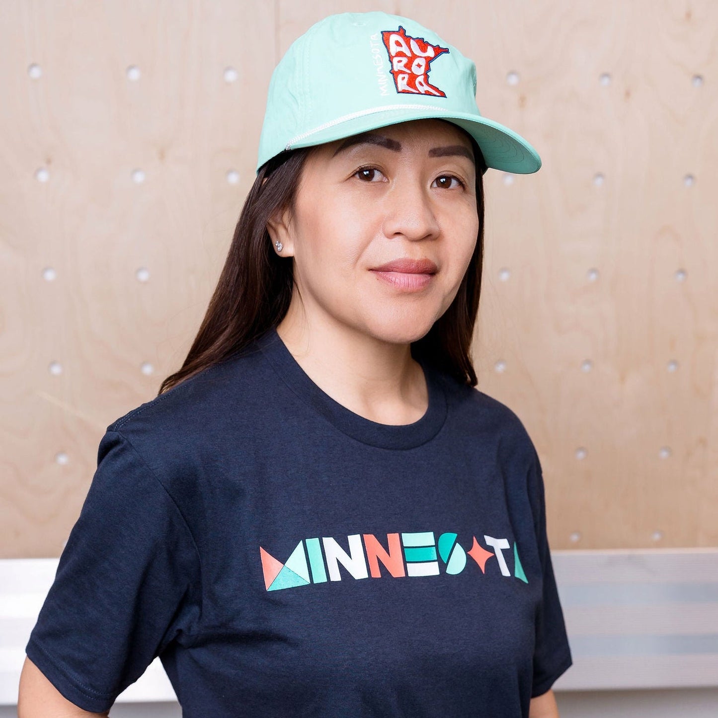 Person wearing a navy blue t-shirt with colorful text and a light blue cap, standing against a beige wall.