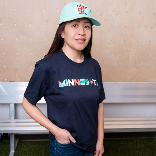 Person wearing a navy 'MINNESOTA' t-shirt and light blue cap indoors.
