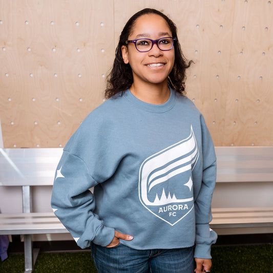 Person wearing a blue sweatshirt with a logo, standing in front of a beige wall and bench.