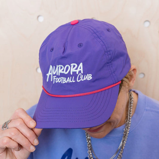 Person wearing a purple 'Aurora Football Club' cap and shirt against a wooden background