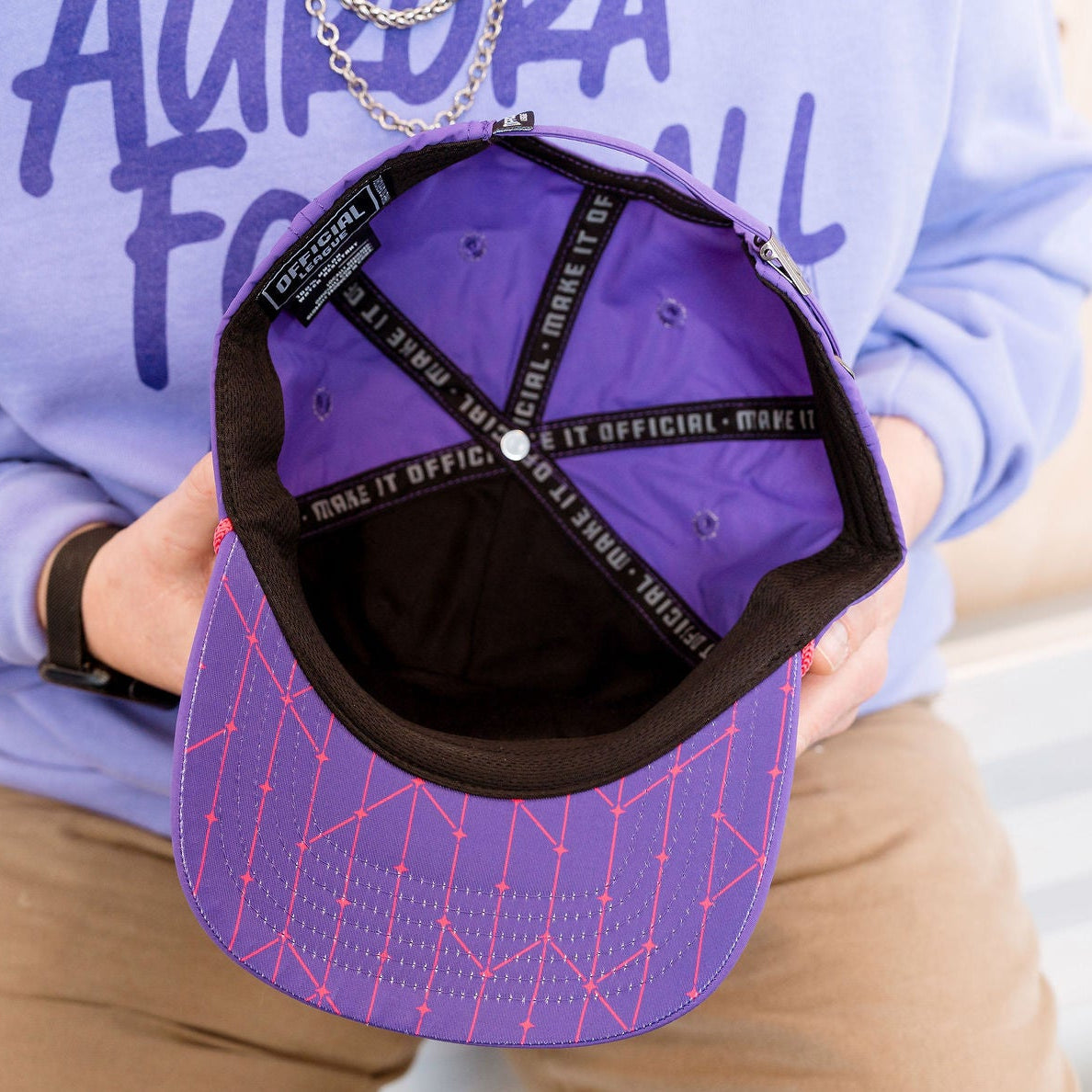 Person holding a purple cap with a geometric design, wearing a light blue sweatshirt with text.