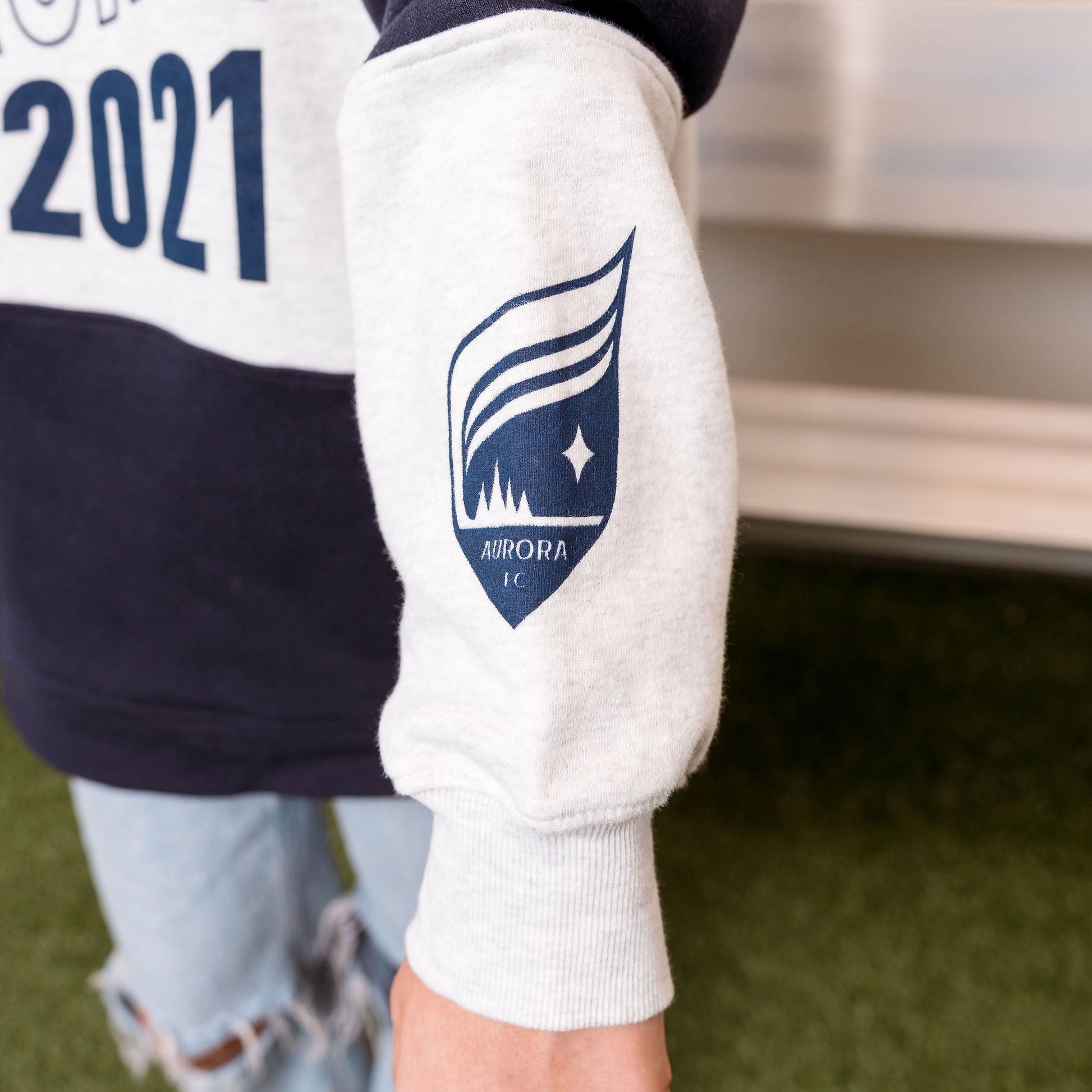 Person wearing a sweatshirt with 'Aurora 2021' text and a logo on the sleeve, standing on grass.