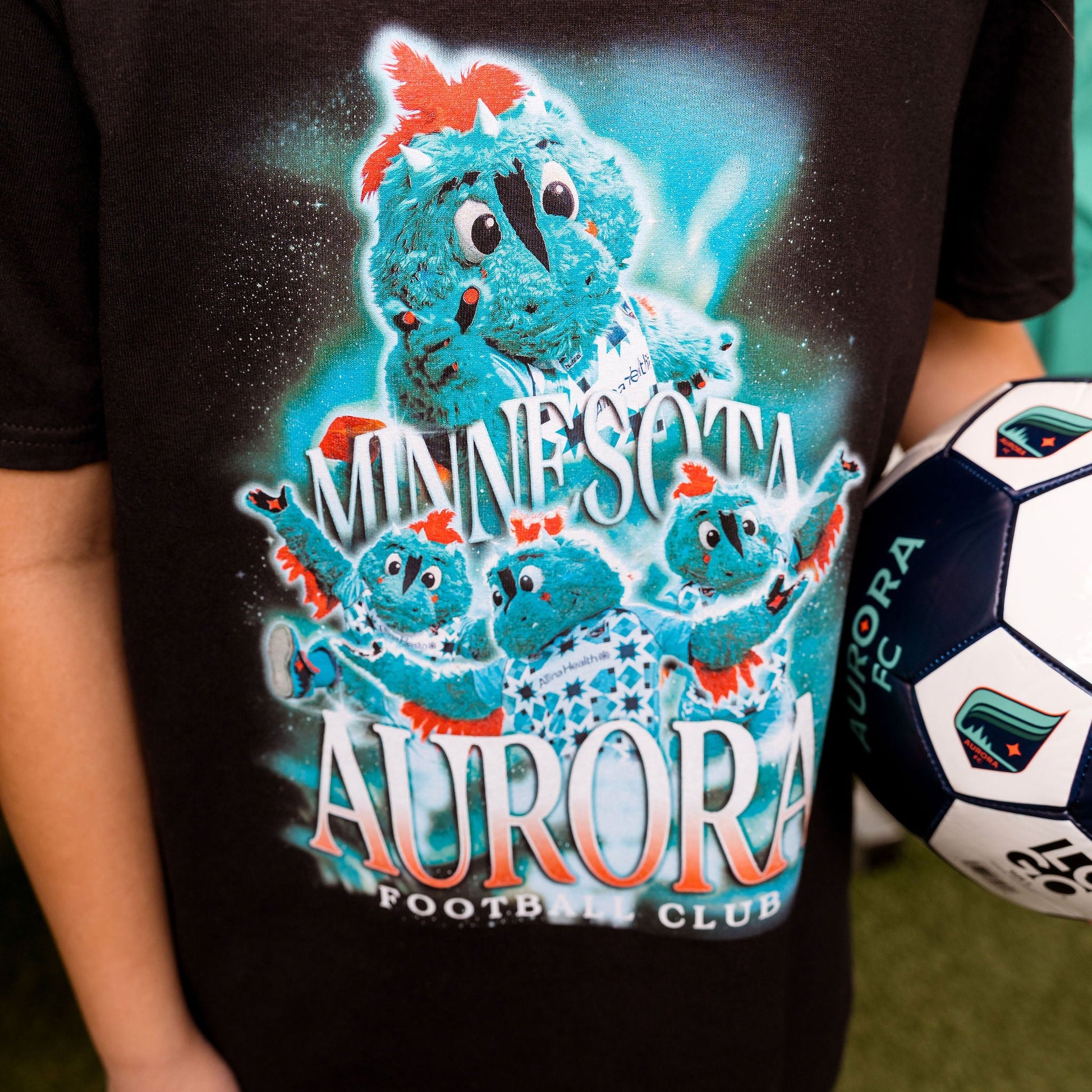 Person wearing a black t-shirt with a colorful graphic of a Rory and 'Minnesota Aurora Football Club' text, holding a soccer ball.