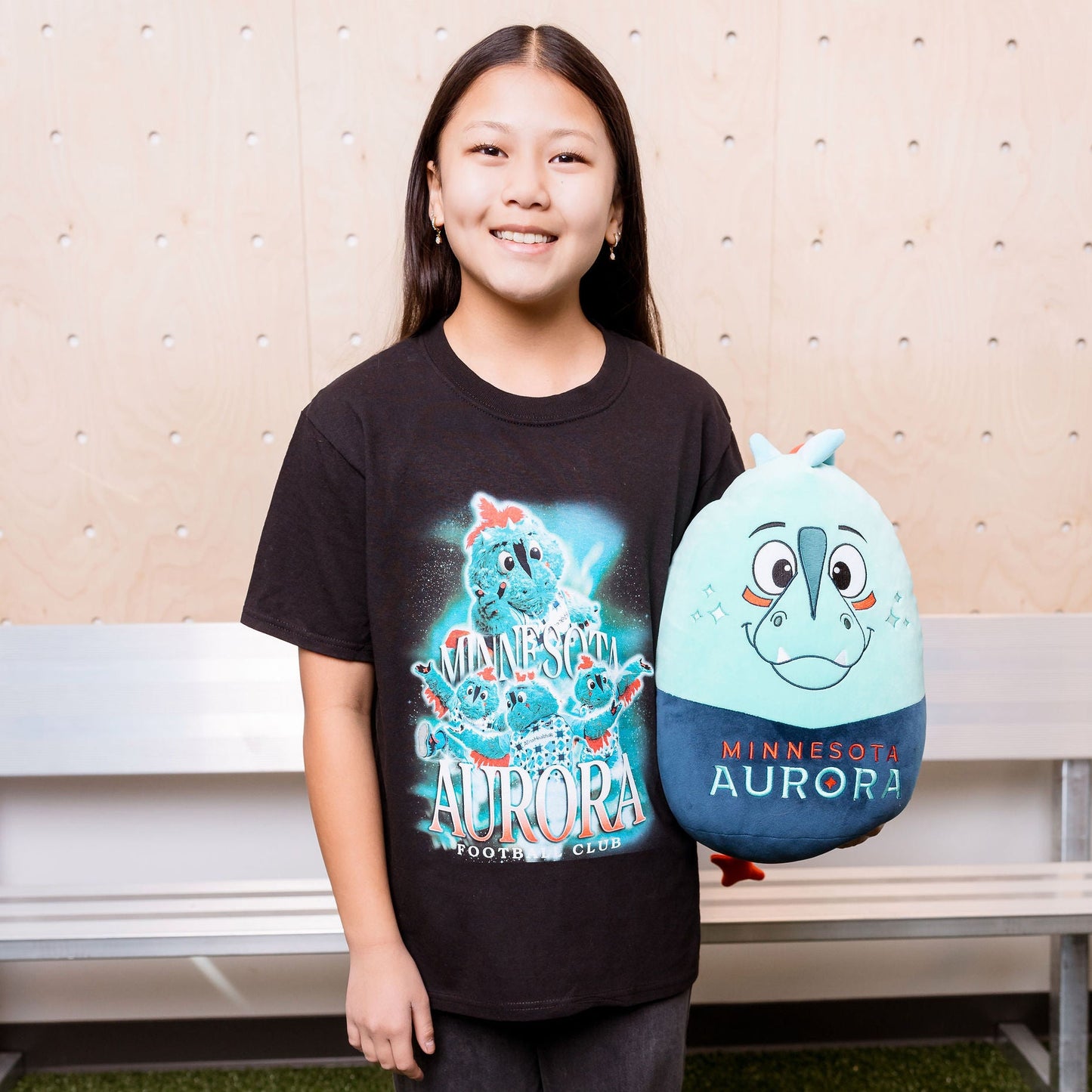 Person wearing a black t-shirt with a colorful design, holding a blue plush toy, standing on grass with a bench and wall in the background.